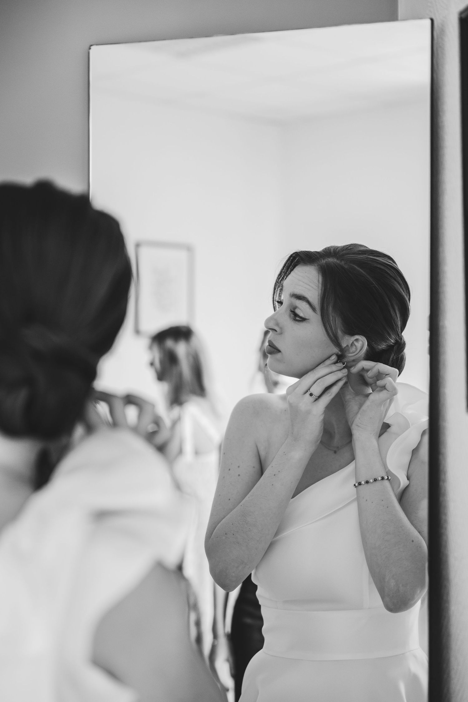 Black and white photo of a bride putting on earrings while looking in a full-length mirror on her wedding day.