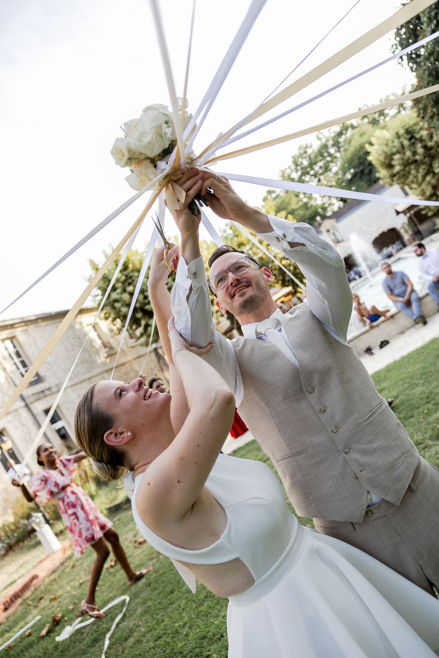 Bride and groom joyfully holding a decorated wedding bouquet with ribbons outdoors on a sunny day.