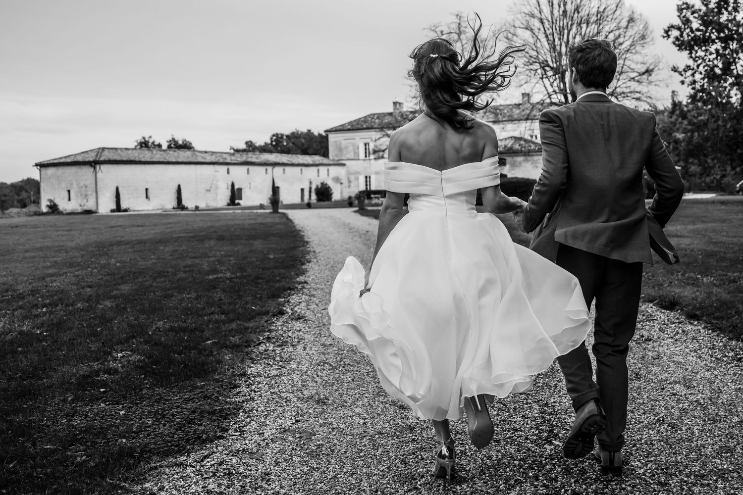 Black and white photo of bride and groom walking hand in hand toward an elegant estate on their wedding day.