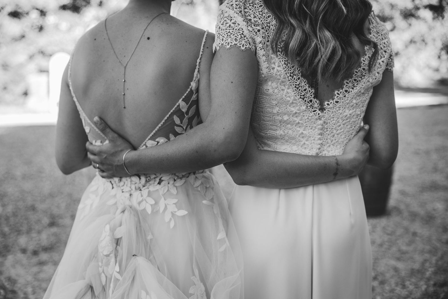 Two brides in elegant white wedding gowns embrace outdoors in a black and white photo.