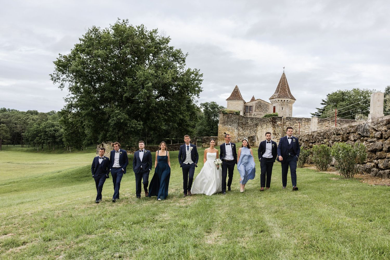 Wedding party posing on green lawn before a historic stone castle on a cloudy day.