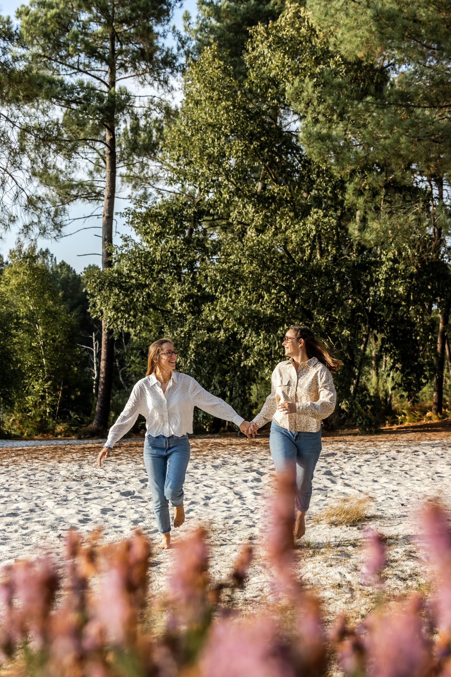 Two women holding hands and running on a sandy beach path surrounded by lush green pine trees and pink wildflowers.
