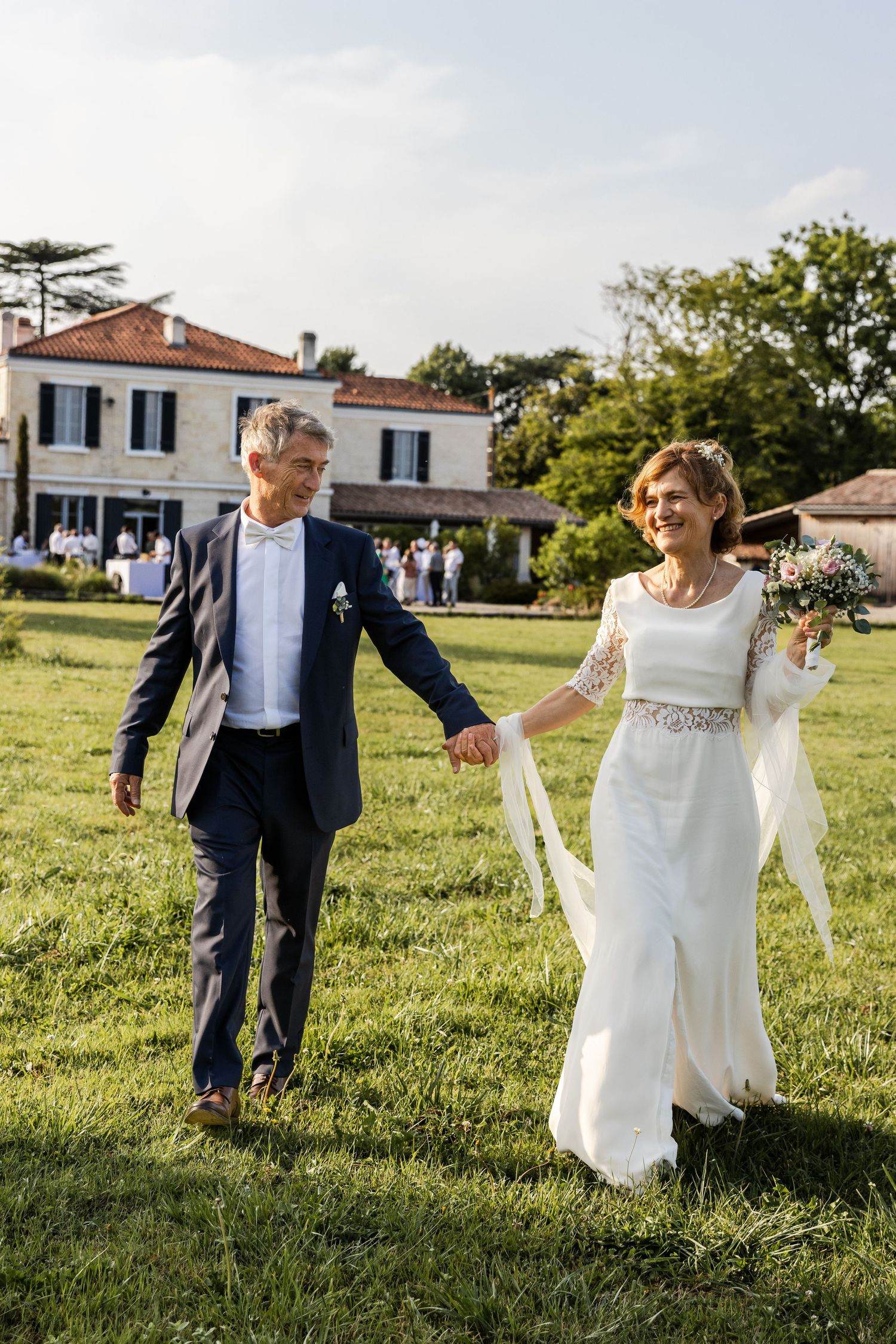 Mature bride and groom holding hands walking on green lawn outside elegant villa during outdoor wedding celebration.