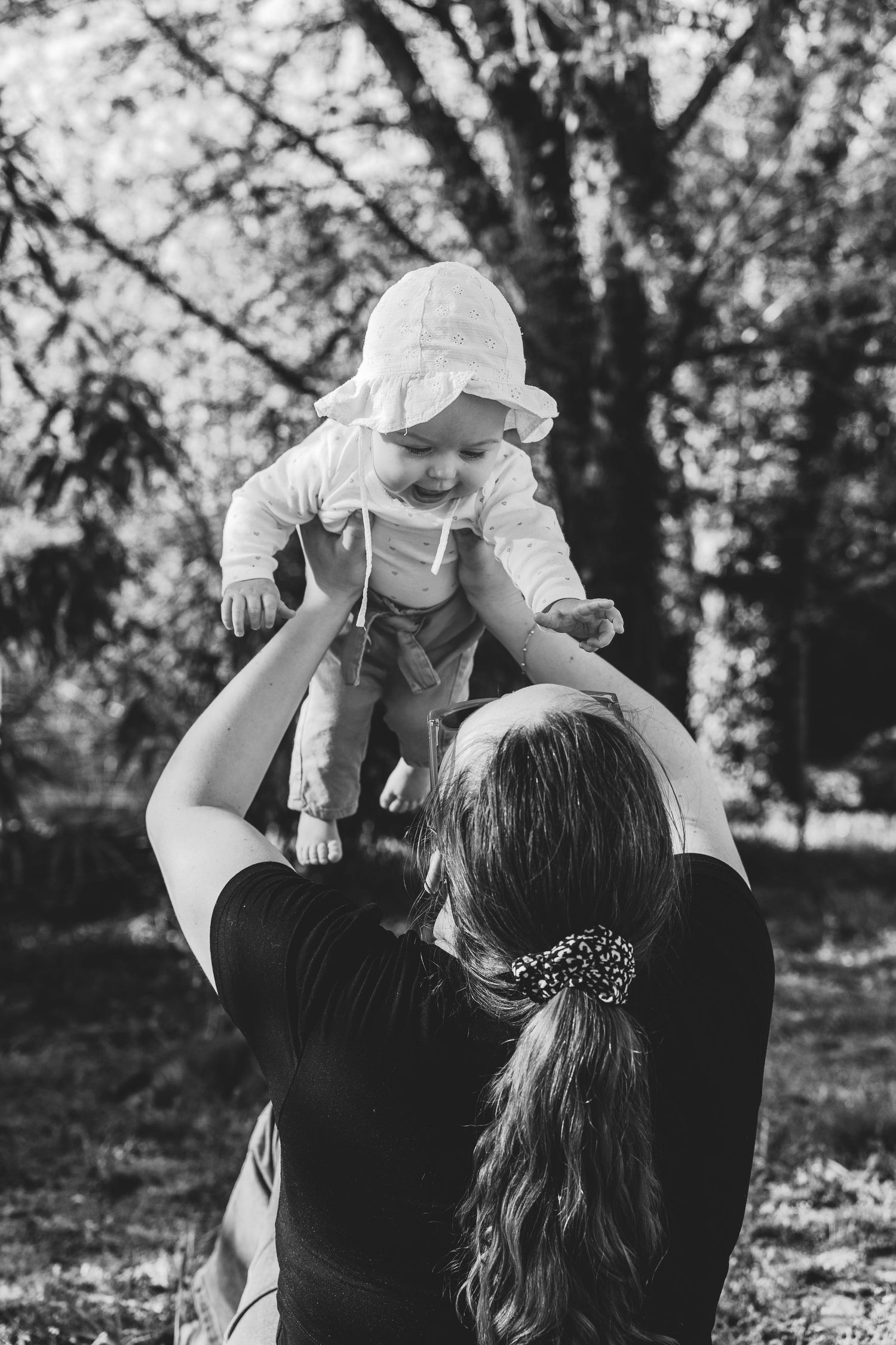Black and white photo of a mother lifting her smiling baby in a white bonnet outdoors in a lush green park.