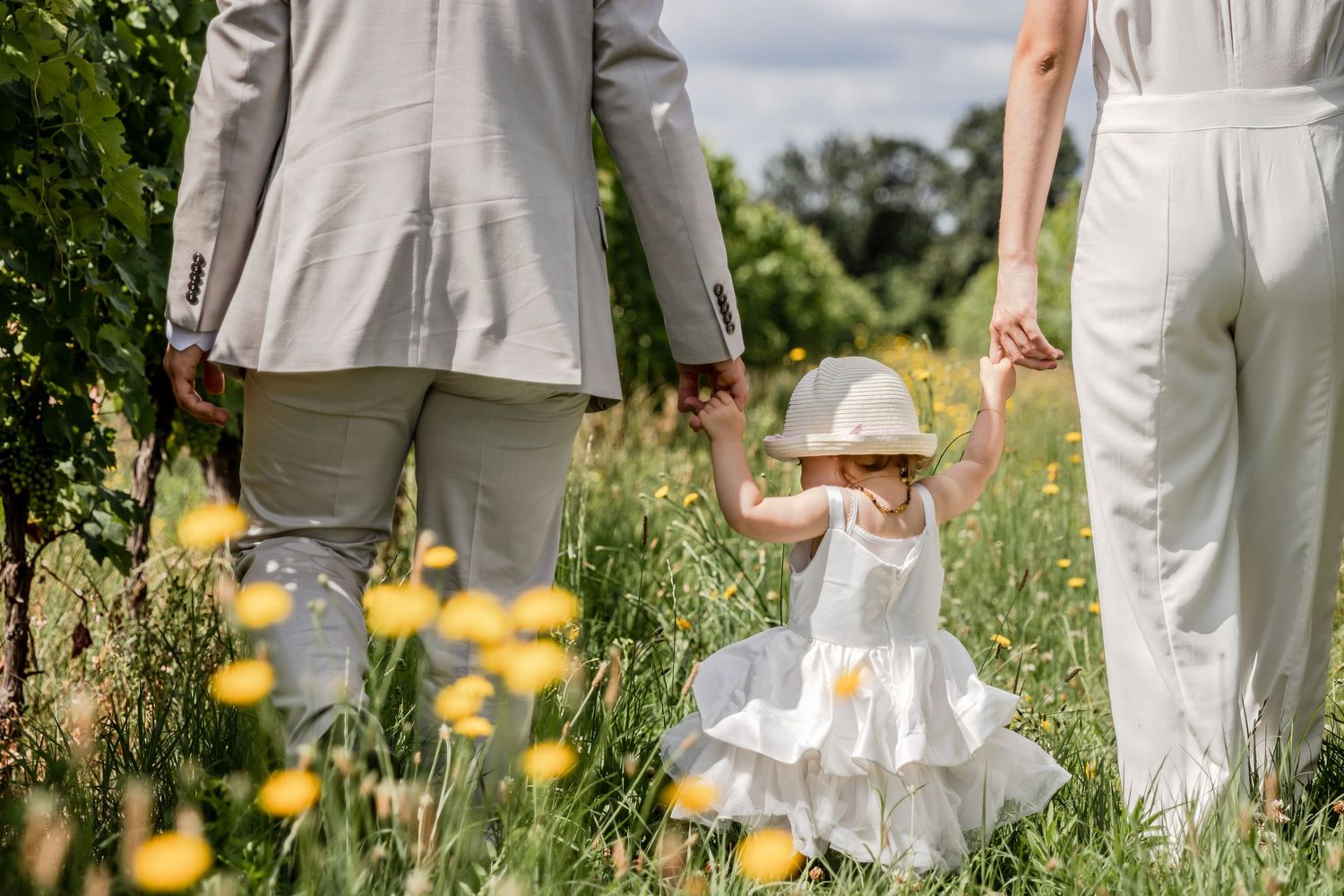A young girl in a white dress and hat holds hands with her parents while walking through a sunny green meadow.