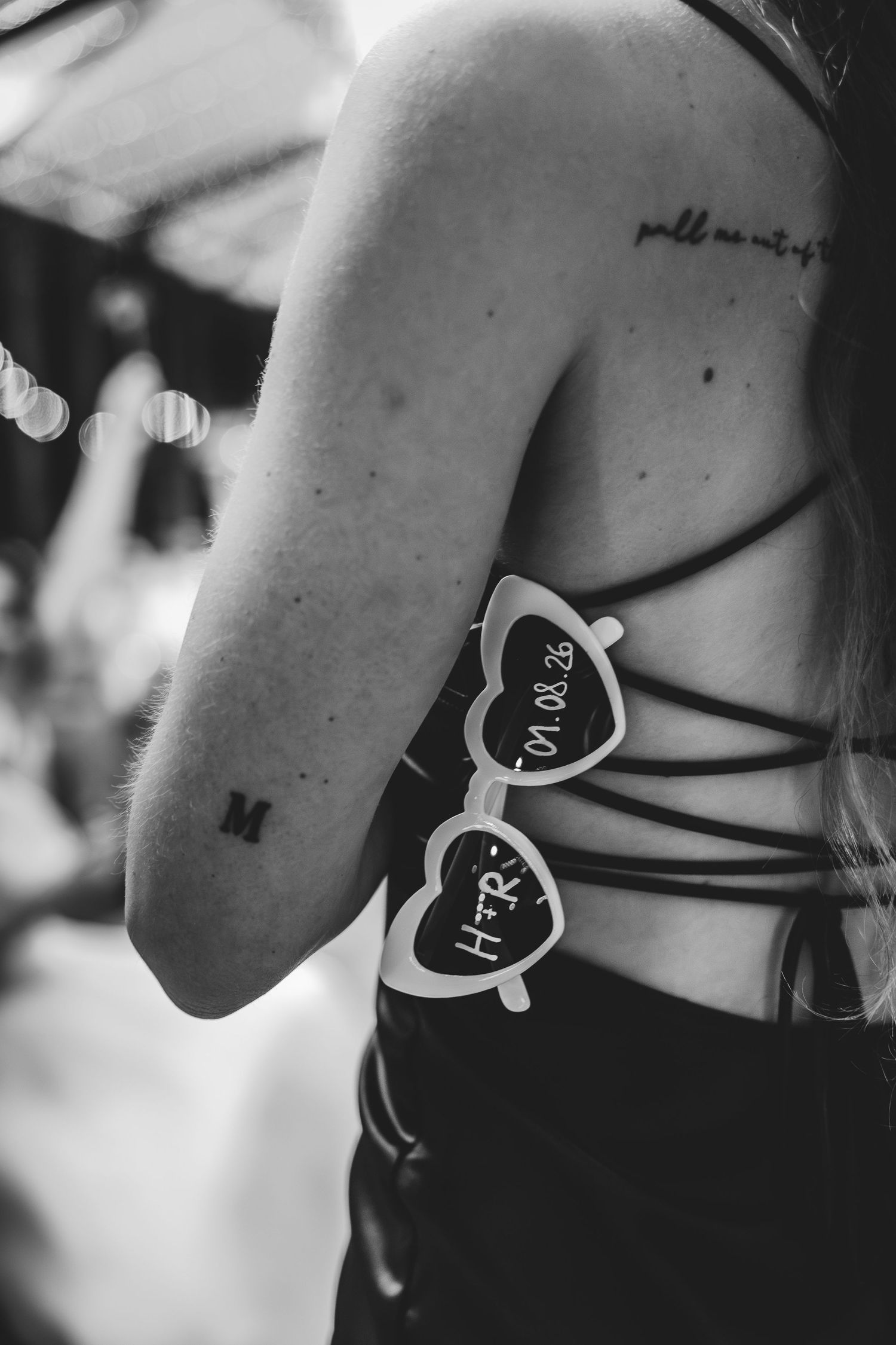 Black and white photo of a woman's back with tattoos and heart-shaped sunglasses tucked into her strappy top.