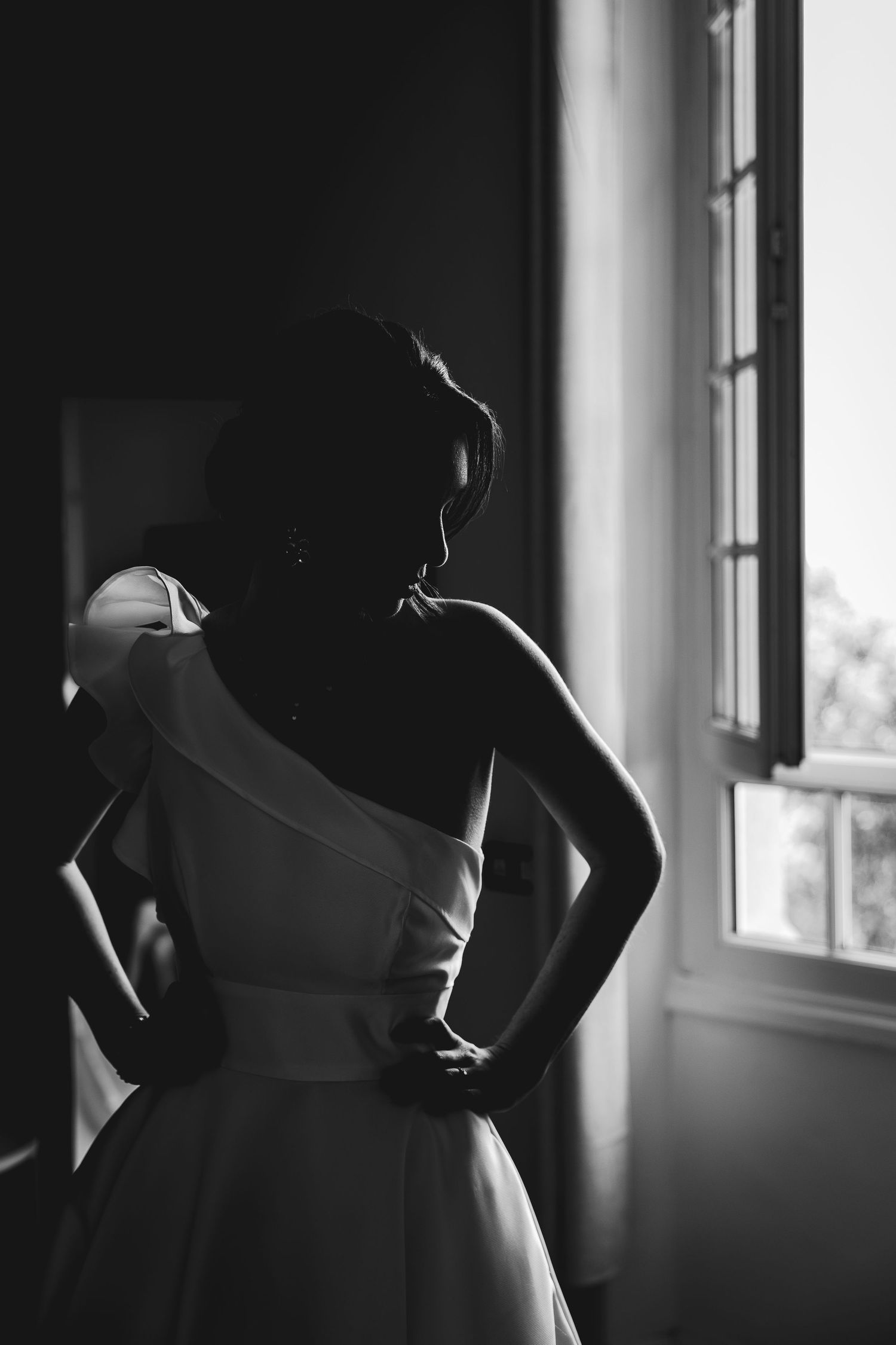 Elegant bride in one-shoulder wedding gown stands by window in black and white portrait photo.