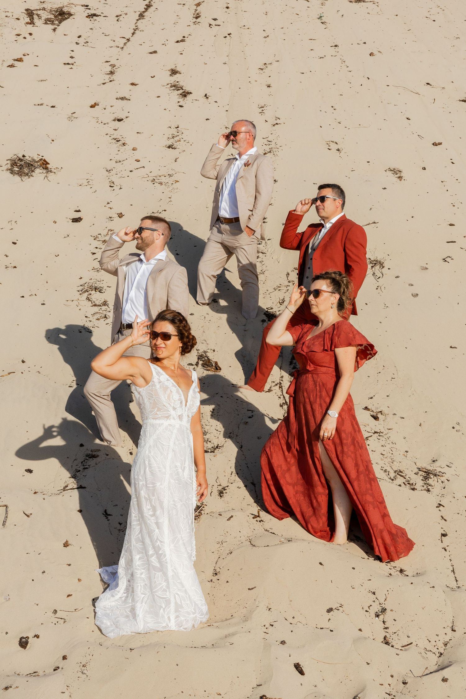 Aerial view of wedding party in formal attire on sandy beach, bride in white gown with guests in tan and red outfits.