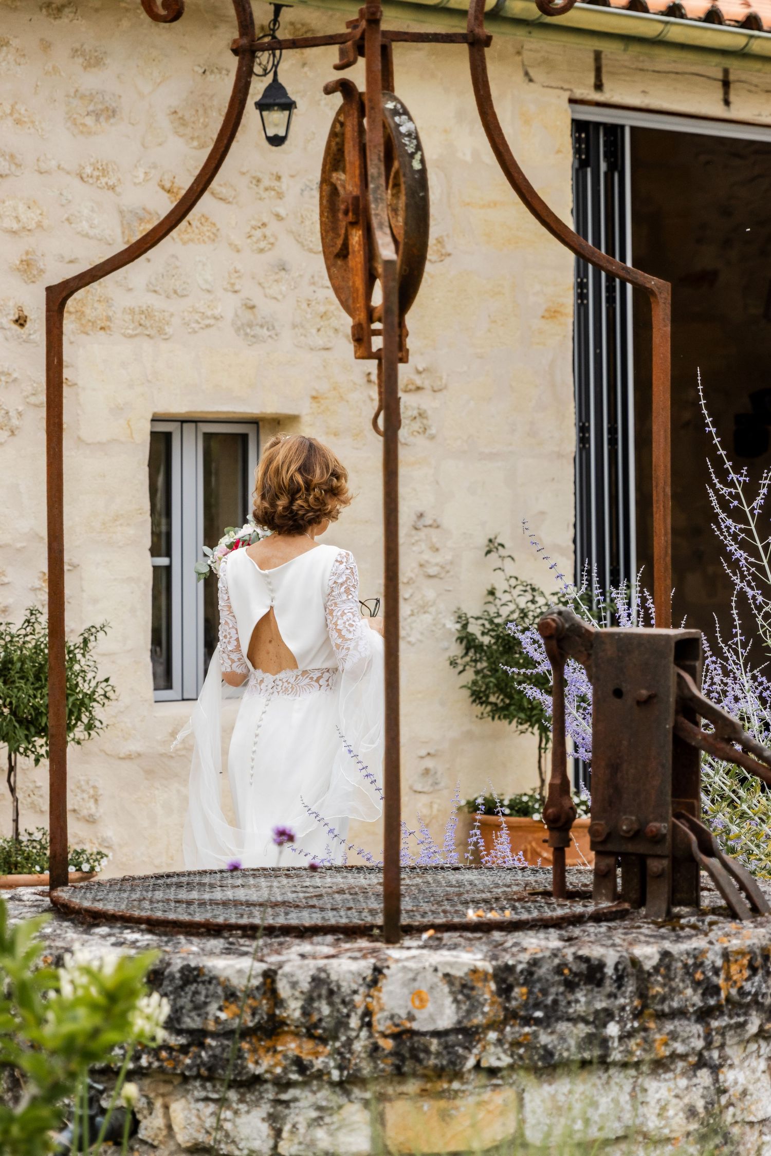 Bride in white lace wedding dress standing near rustic stone well at vintage countryside venue.
