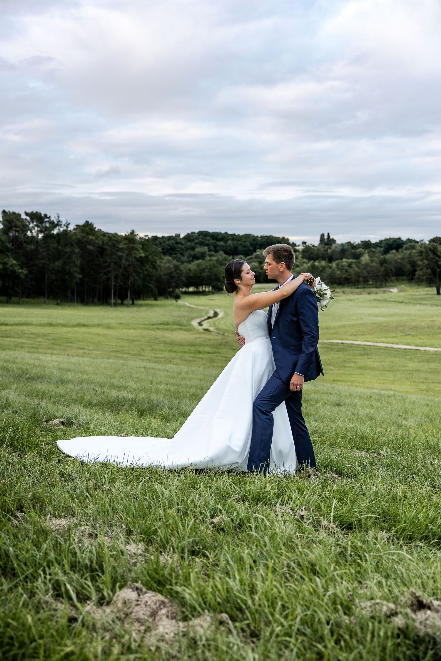 Bride and groom embracing in a lush green field, bride in white gown with train, groom in navy suit.