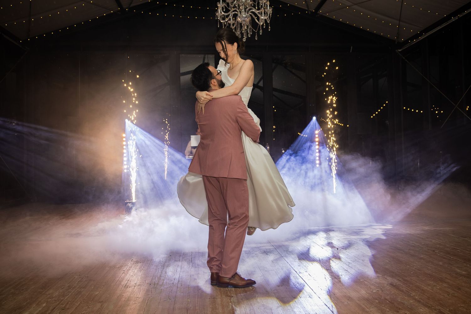 Groom in pink suit lifts bride in white dress during first dance on foggy dance floor with sparkling lights.