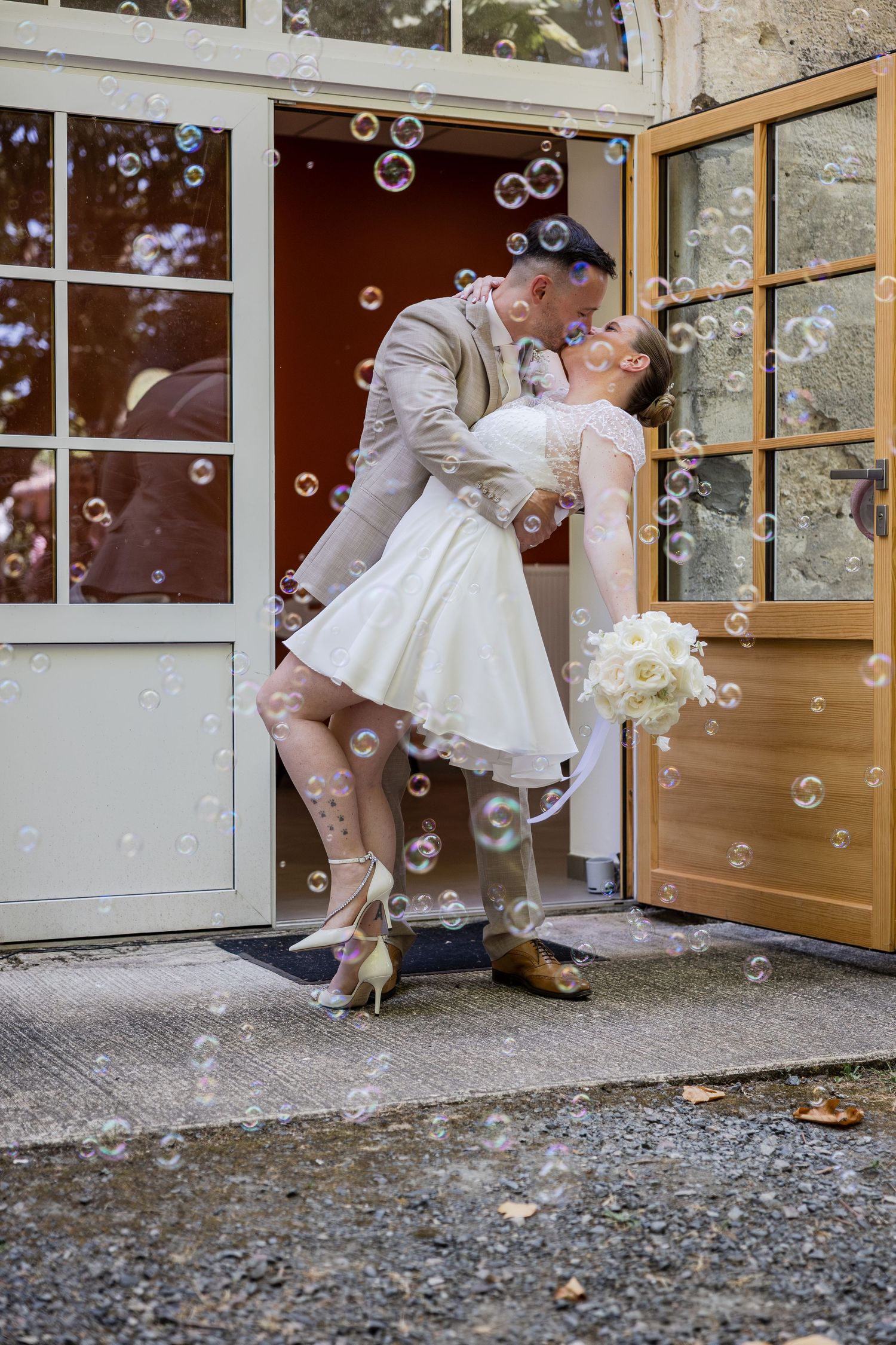 Bride and groom share a romantic dip kiss in a doorway surrounded by floating soap bubbles on their wedding day.