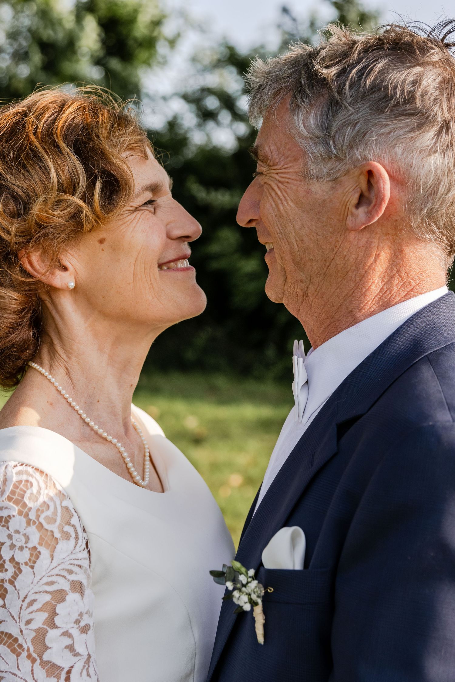 Smiling mature couple in wedding attire gazing lovingly at each other outdoors, woman in lace dress with pearl necklace.