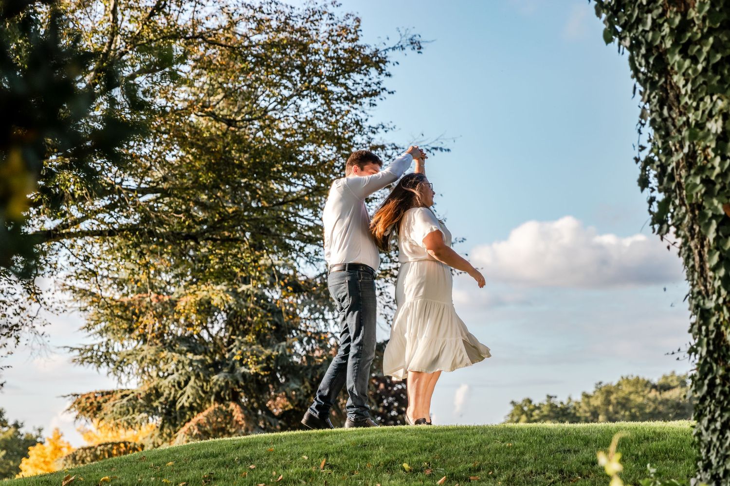 A couple dances together on a lush green hill surrounded by trees on a sunny day.