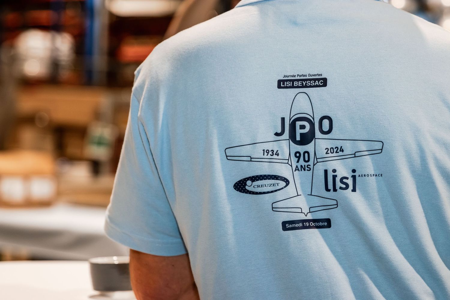 Light blue t-shirt with JPO 90 ans anniversary graphic featuring a vintage airplane design, worn by a person indoors.