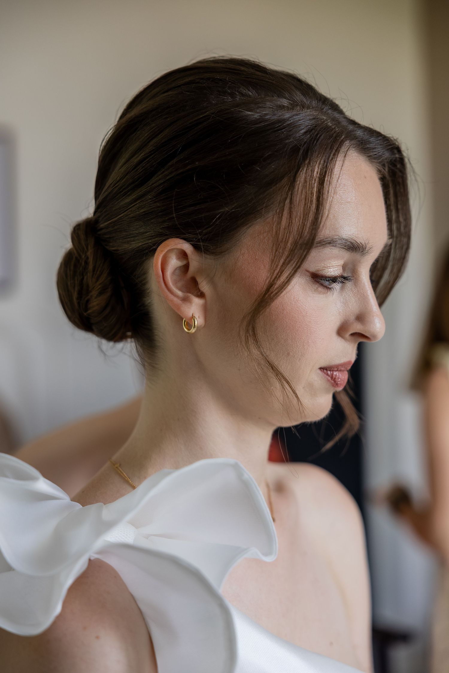 Bride with elegant updo hairstyle and gold hoop earrings wearing a white off-shoulder gown with bow detail.