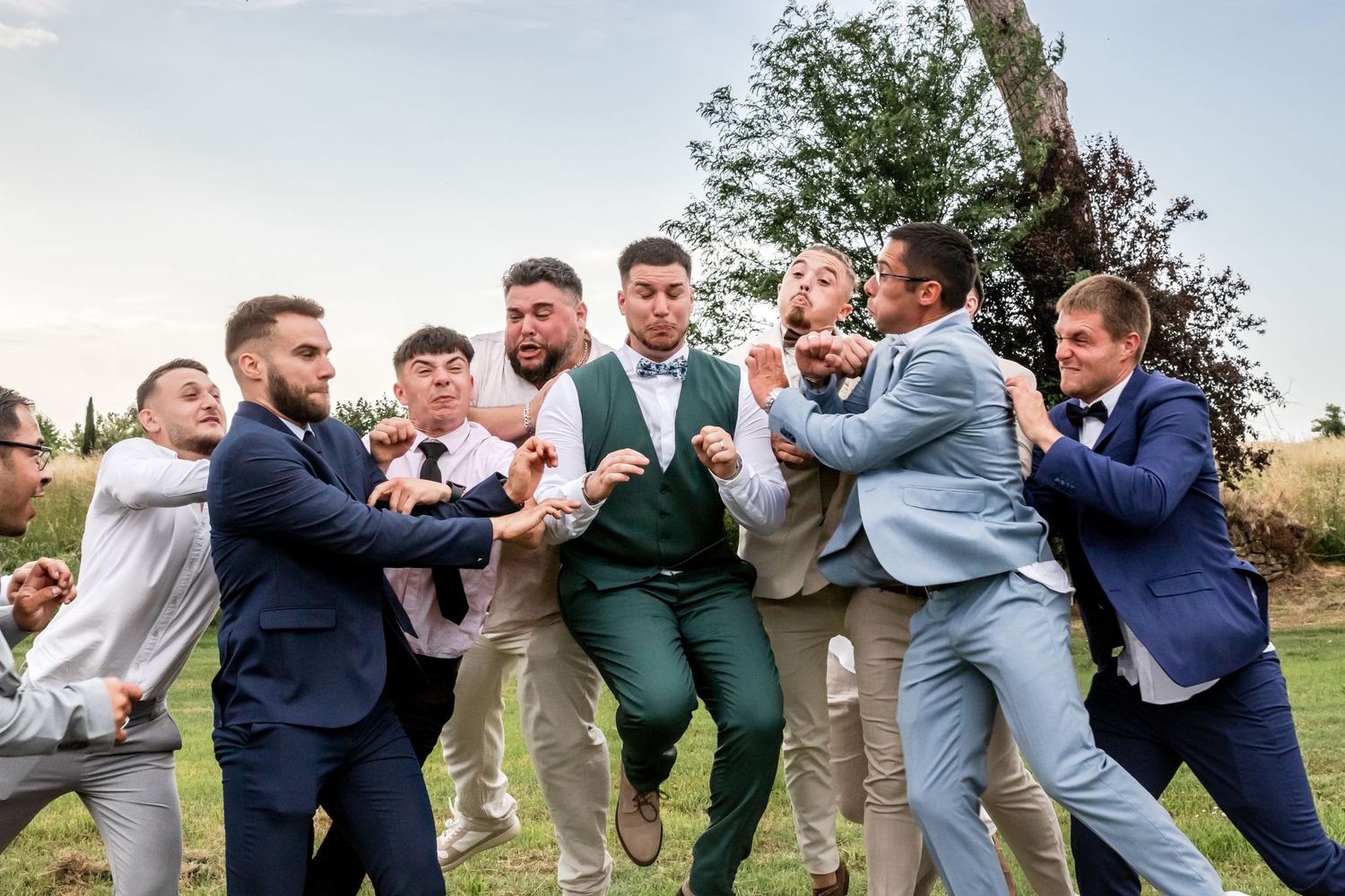 Groom in green suit playfully pulled by groomsmen in blue suits outdoors at a wedding celebration.