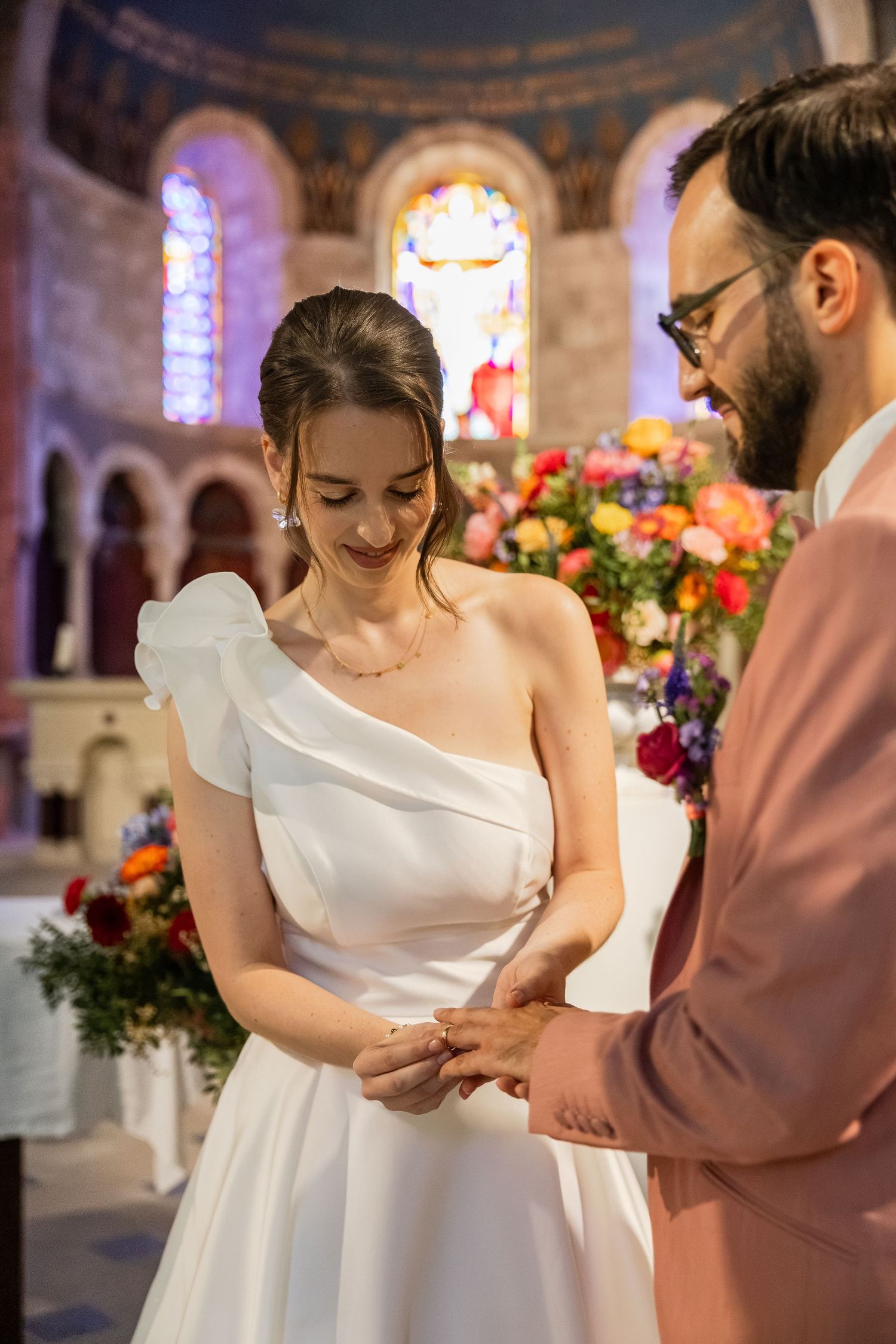 Bride in one-shoulder white gown receives ring from groom in pink suit during church wedding ceremony with floral backdrop.