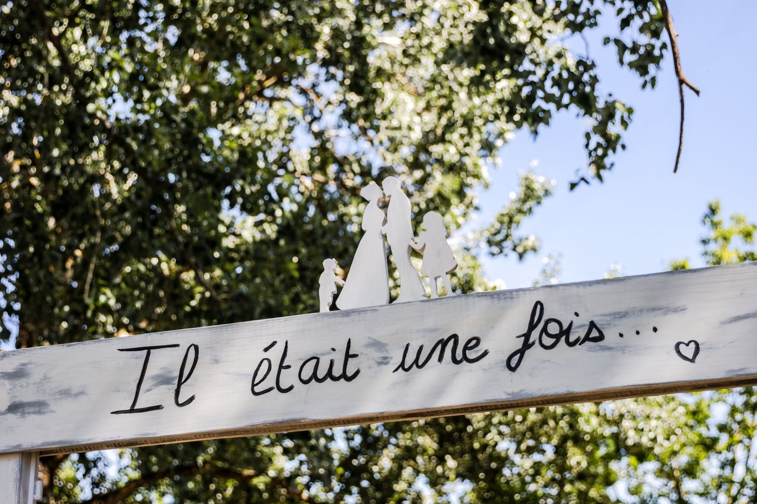 Rustic white wooden sign reading Il était une fois in French script, displayed outdoors with green trees in background.