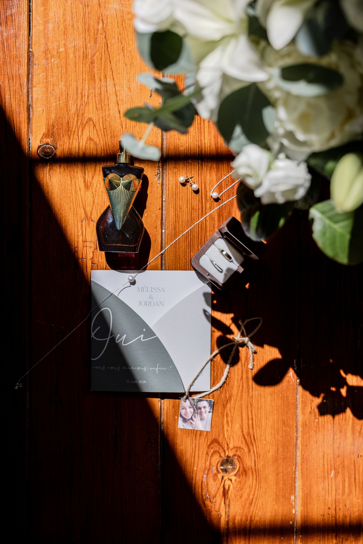 Wedding flat lay with invitation, perfume bottle, earrings, and white floral bouquet on rustic wooden surface.