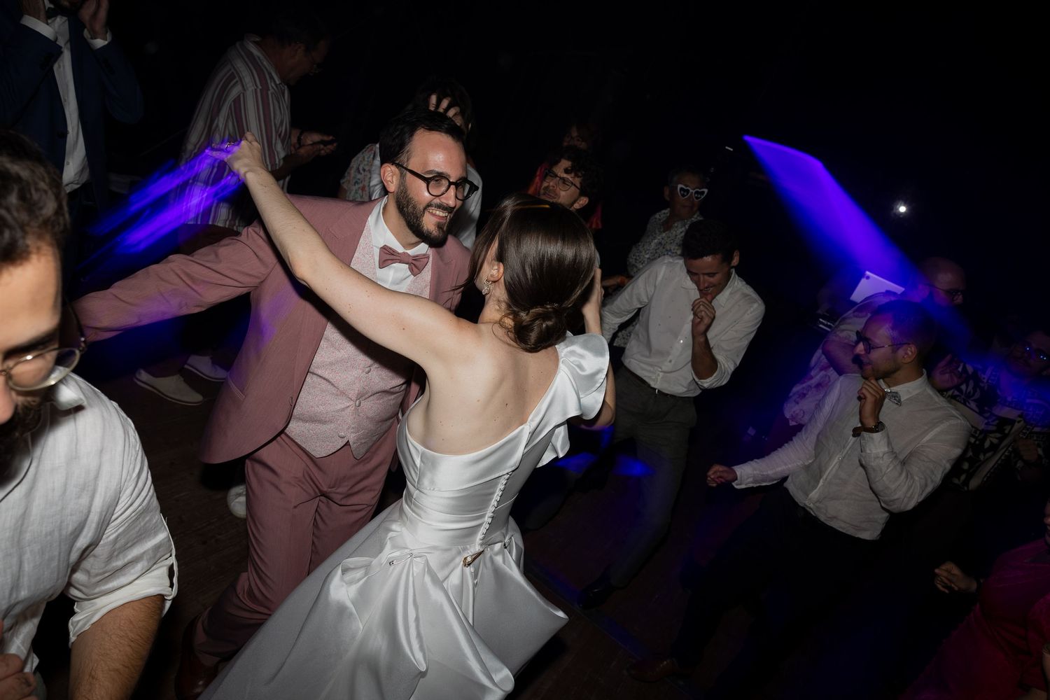 Bride and groom dancing joyfully at wedding reception with colorful blue and purple lights.