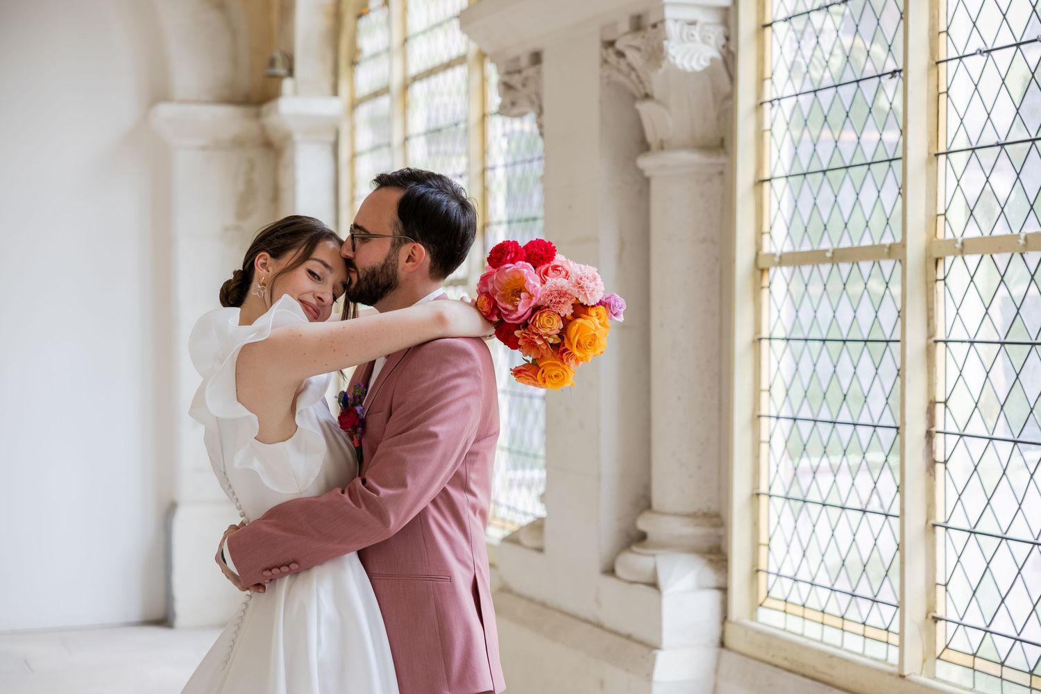 Bride and groom embracing near ornate windows, groom kissing bride's forehead, bride holding colorful pink and orange bouquet.