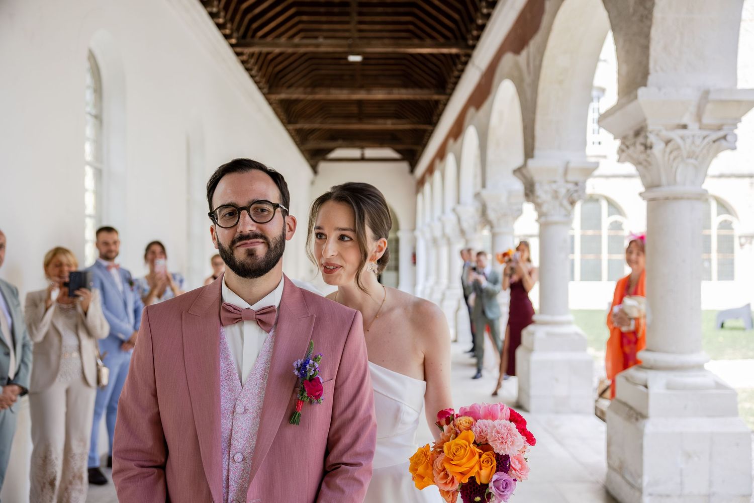 Bride holding colorful bouquet and groom in pink suit with bow tie pose together at elegant wedding venue.