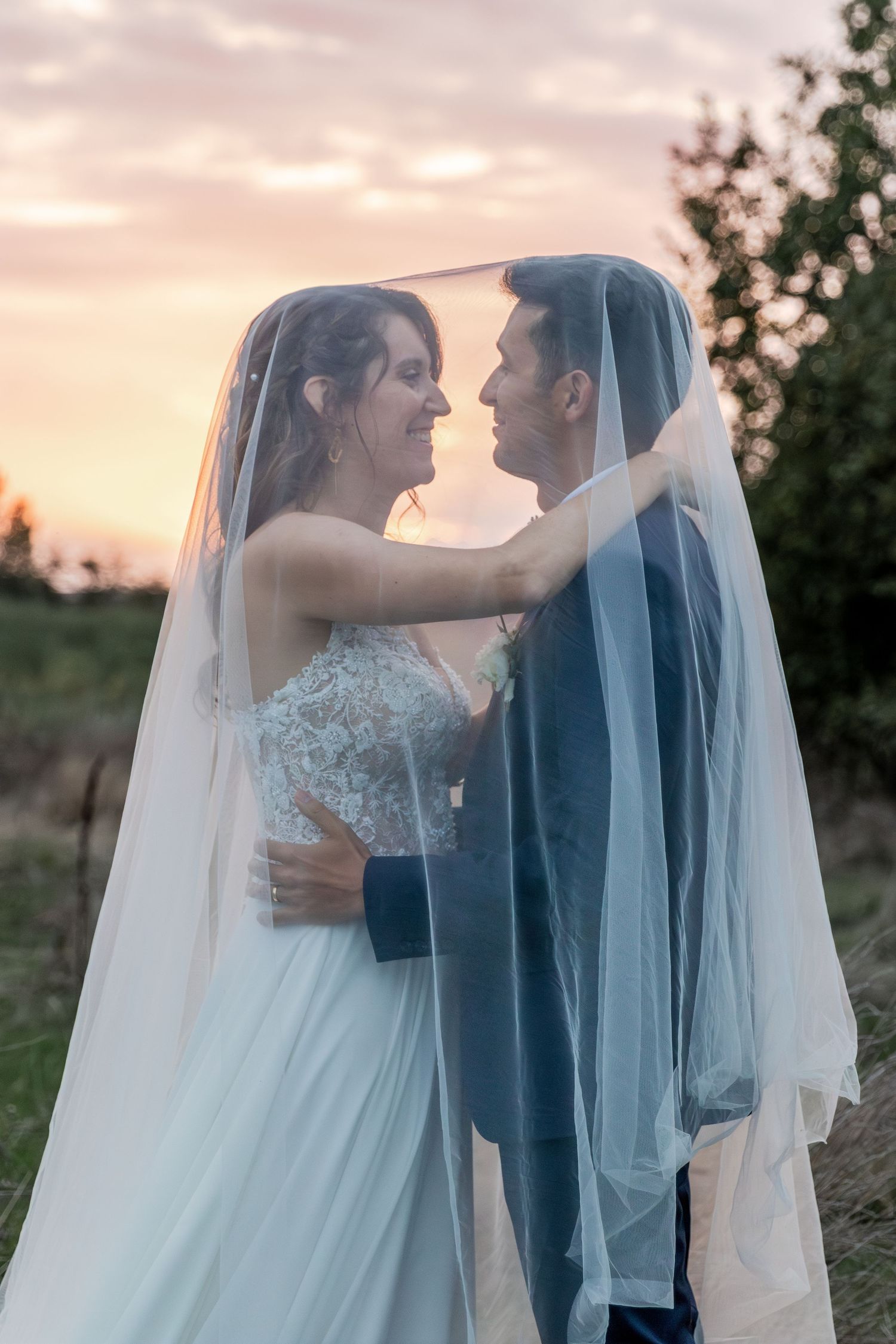 Bride and groom share romantic moment under wedding veil at sunset outdoors.