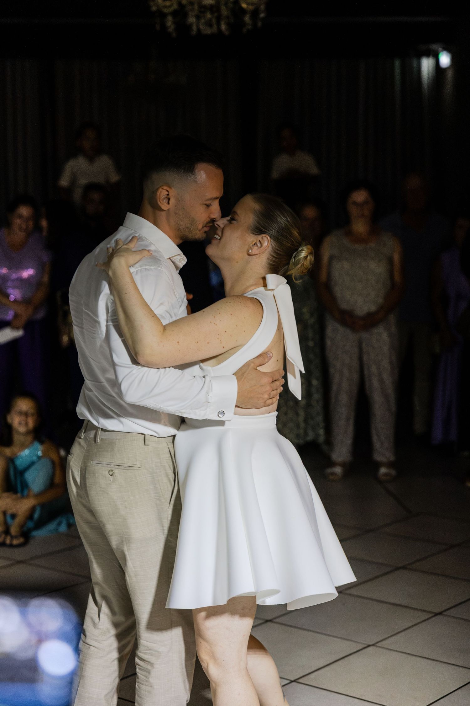 Couple sharing their first dance kiss at wedding reception, bride in white mini dress, guests watching in background.