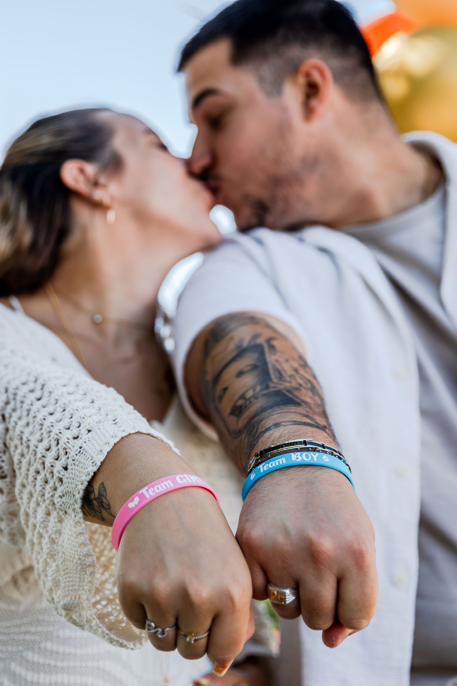 Couple kissing while showing matching pink and blue Team wristbands, man with tattoo sleeve wearing rings.