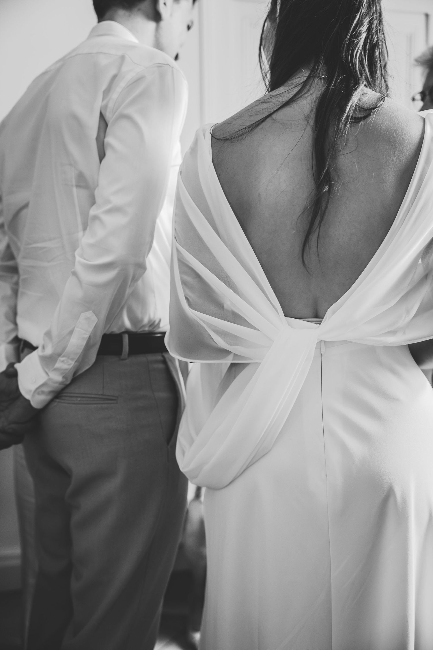 Black and white photo of bride in elegant backless wedding gown standing beside groom in white shirt.