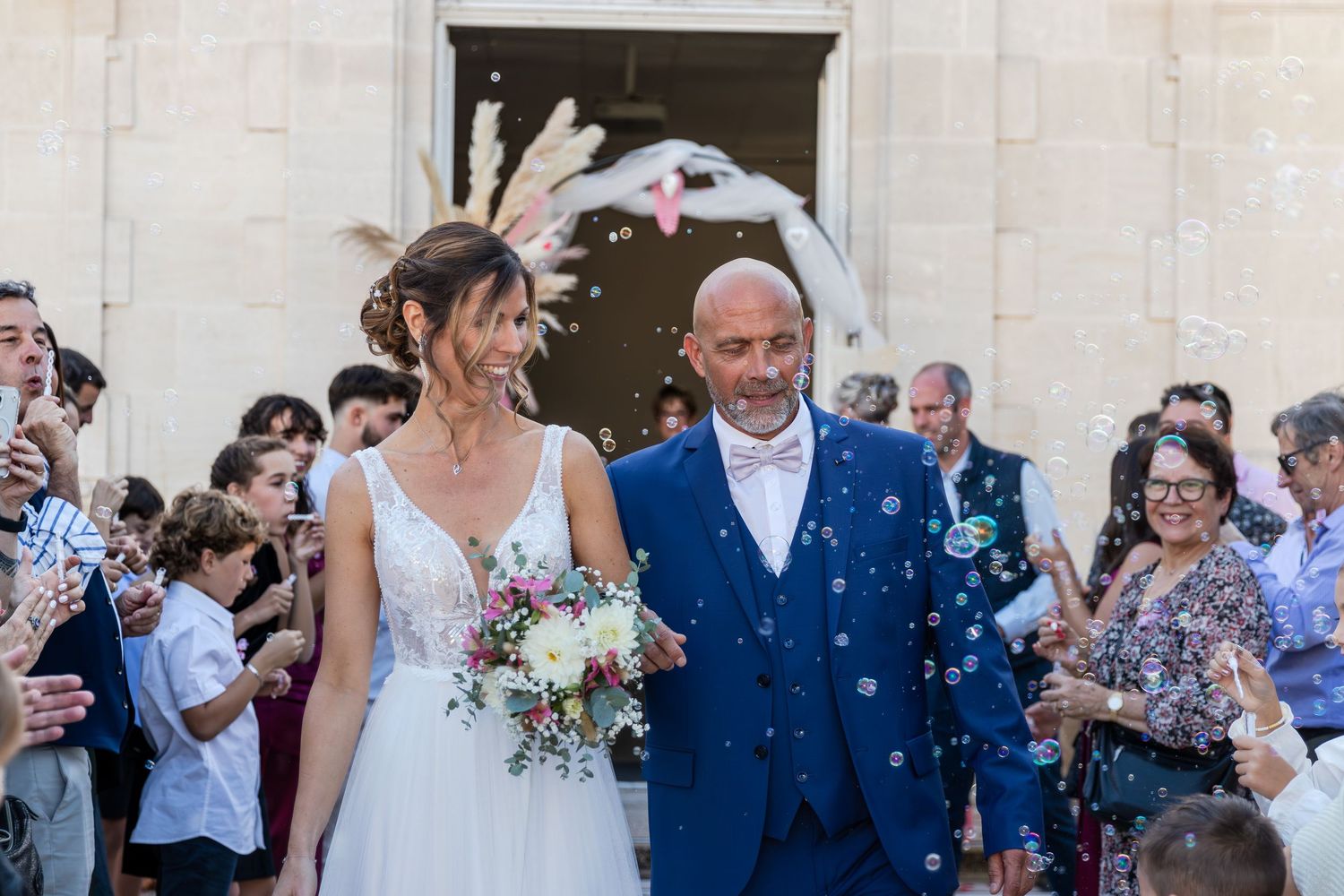 Bride and groom exit church ceremony as guests celebrate with bubbles and confetti outdoors.
