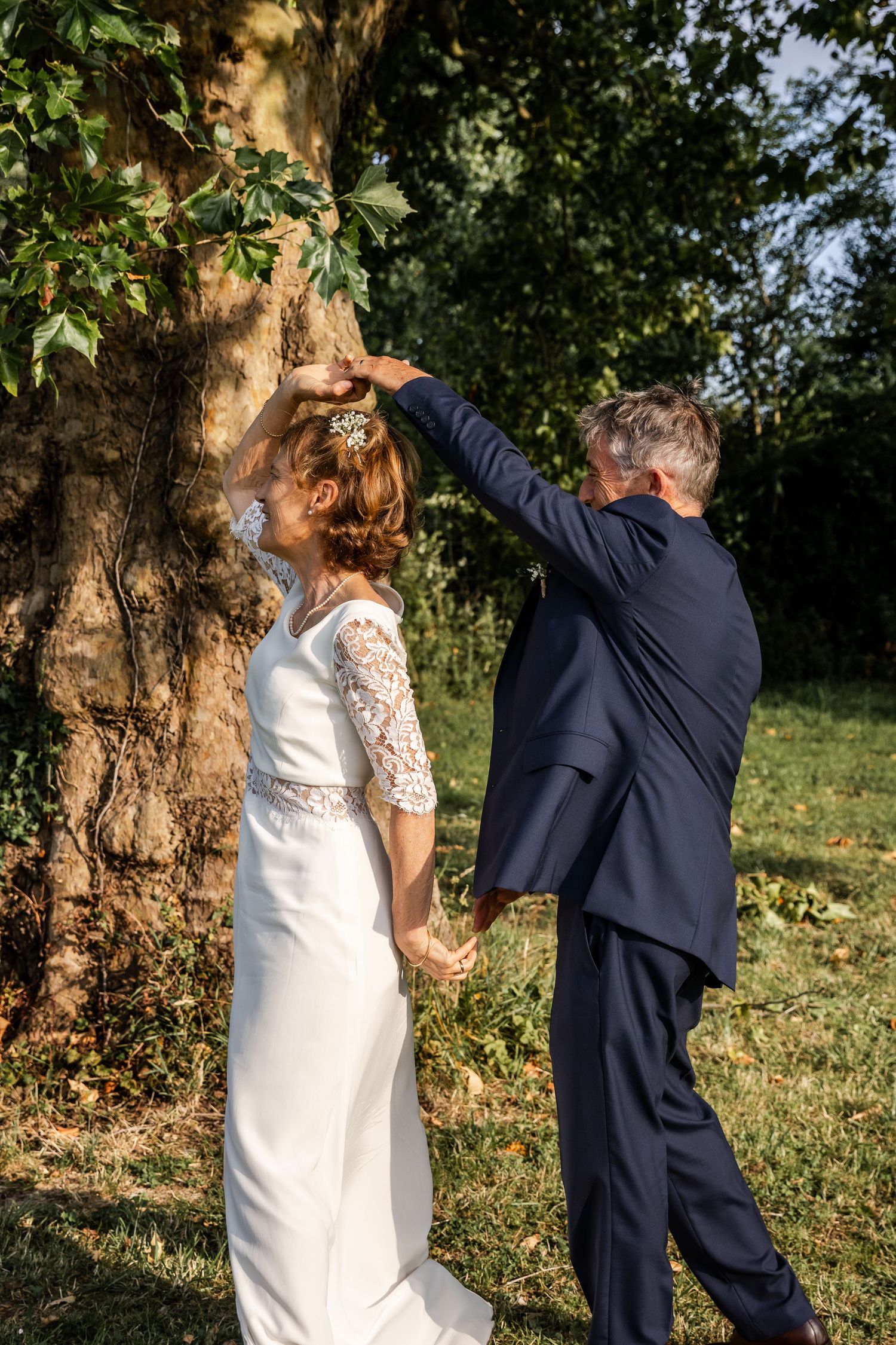 A bride and groom hold hands near a large tree outdoors, the groom raising his arm in a romantic wedding photo.