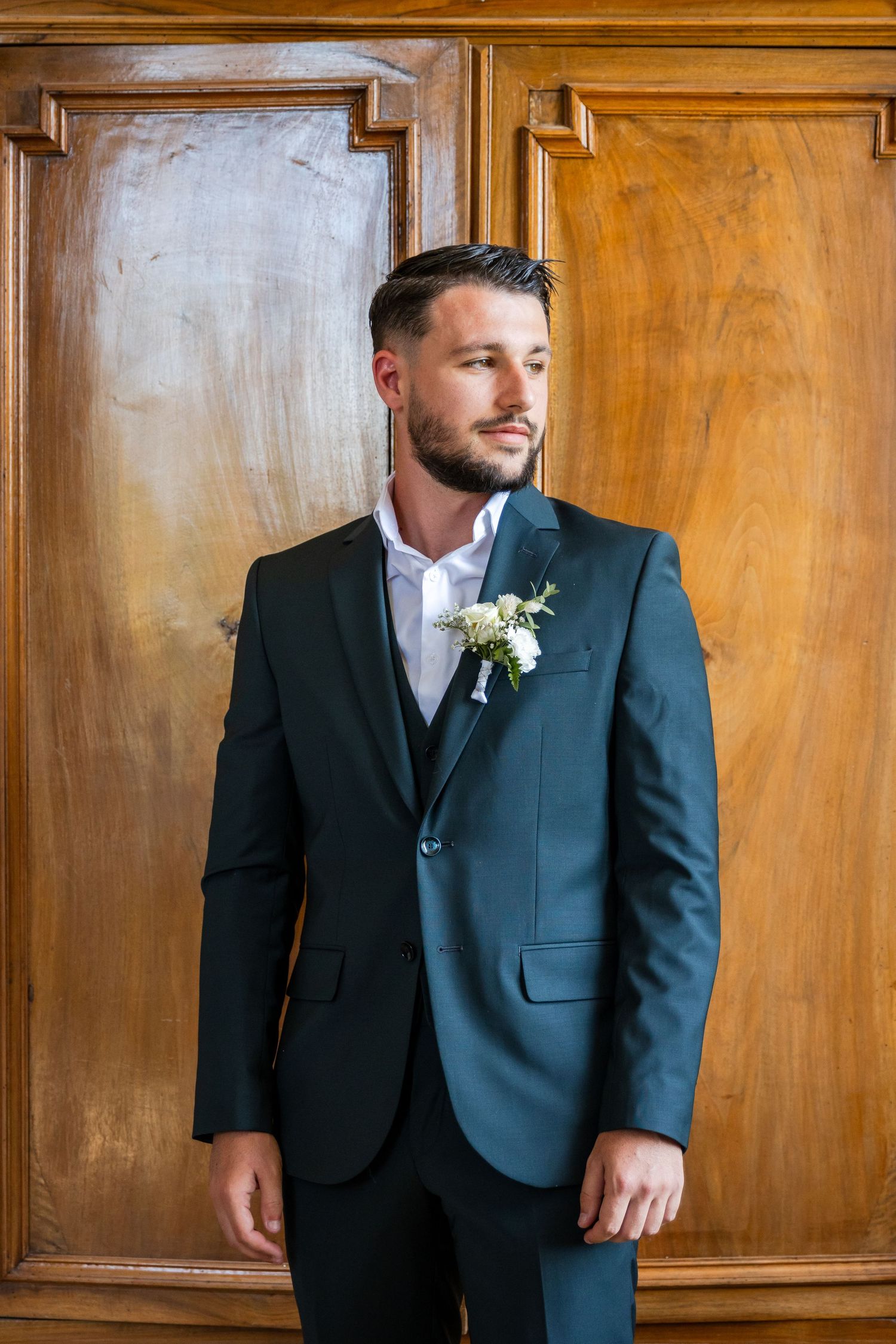 Groom in dark suit with boutonniere stands before wooden doors, looking away thoughtfully.