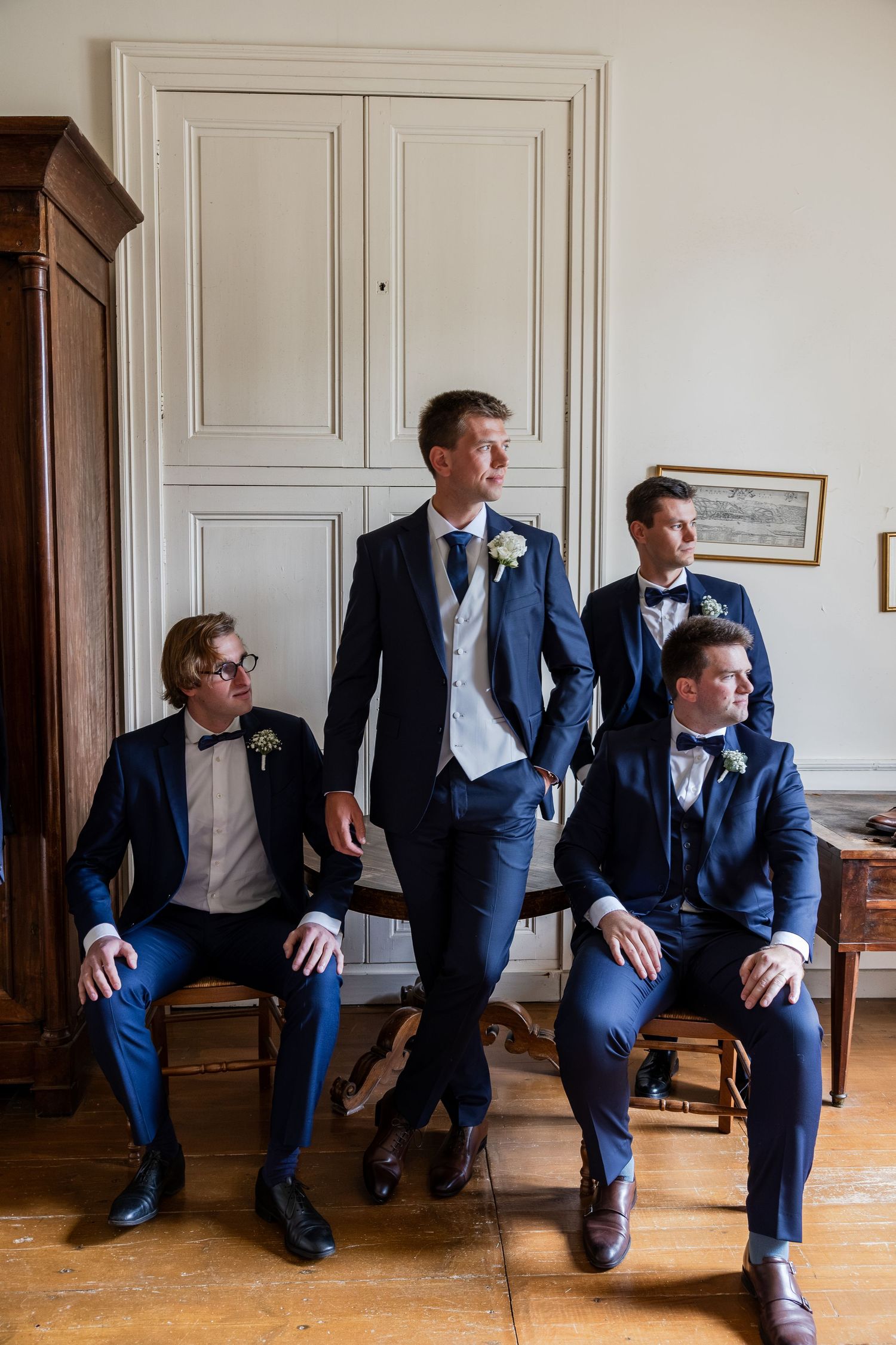 Four groomsmen in navy blue suits with boutonnieres pose elegantly in a classic white-paneled room before a wedding.