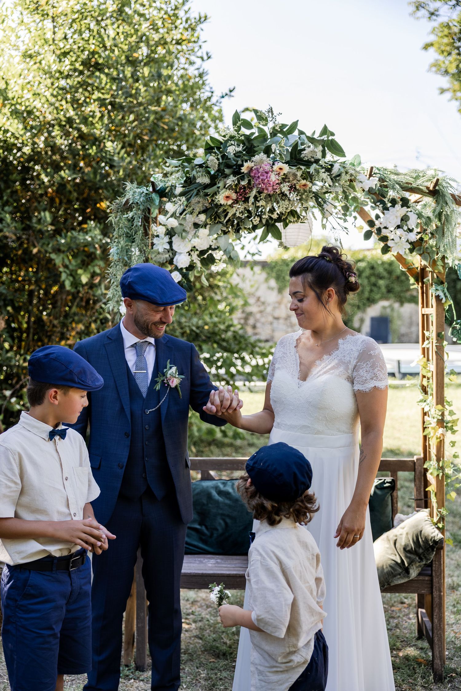 Bride and groom exchange vows outdoors under a floral arch, accompanied by two boys in matching blue caps.