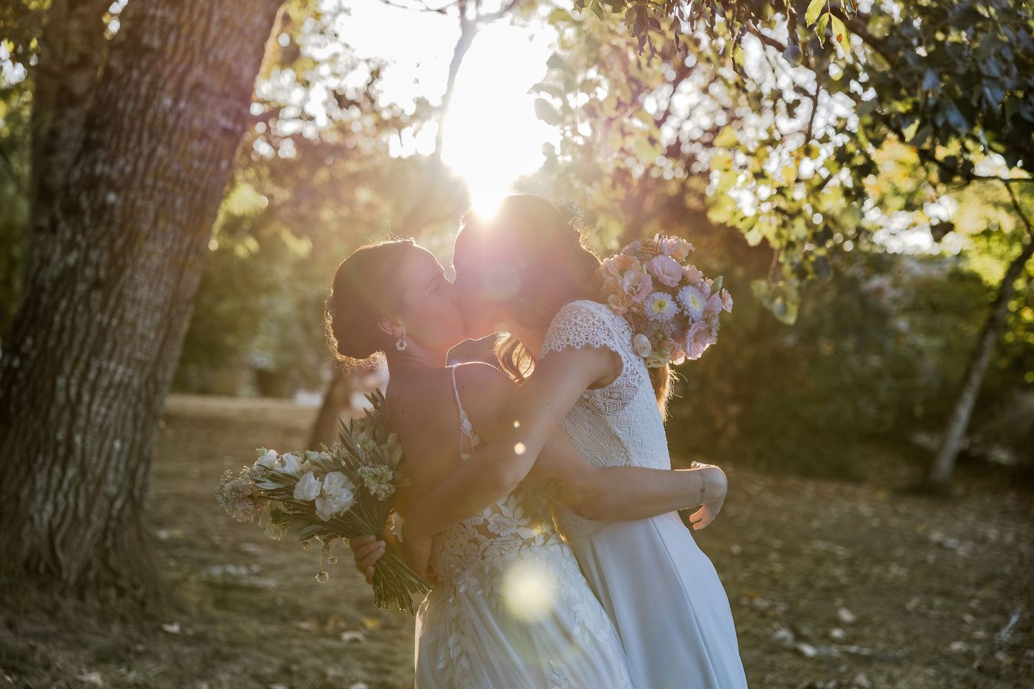 Bride and groom kissing in golden sunlight among trees, holding white floral bouquets in an outdoor wedding setting.