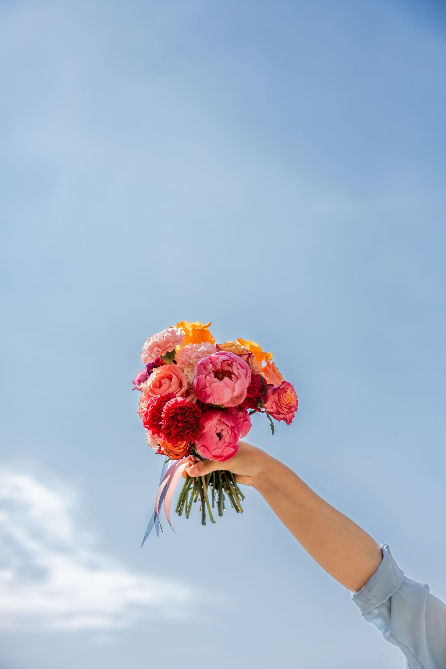 A hand holding a vibrant bouquet of pink, red, and orange ranunculus flowers against a blue sky.