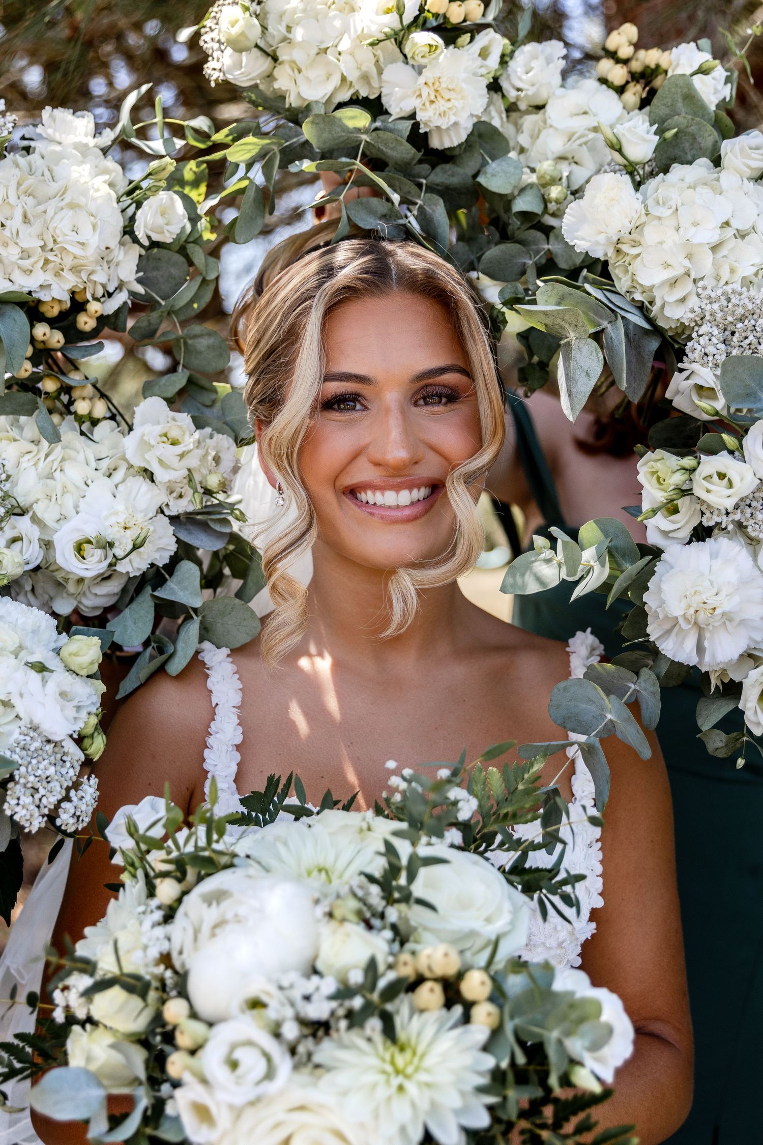Smiling bride holding white floral bouquet surrounded by elegant white roses and eucalyptus greenery arch.