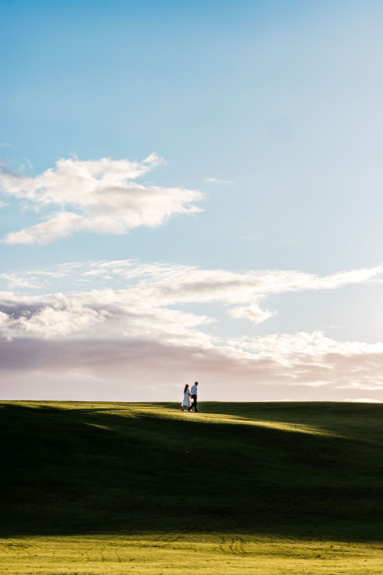 Two people walking on a lush green golf course hill under a bright blue sky with scattered white clouds.