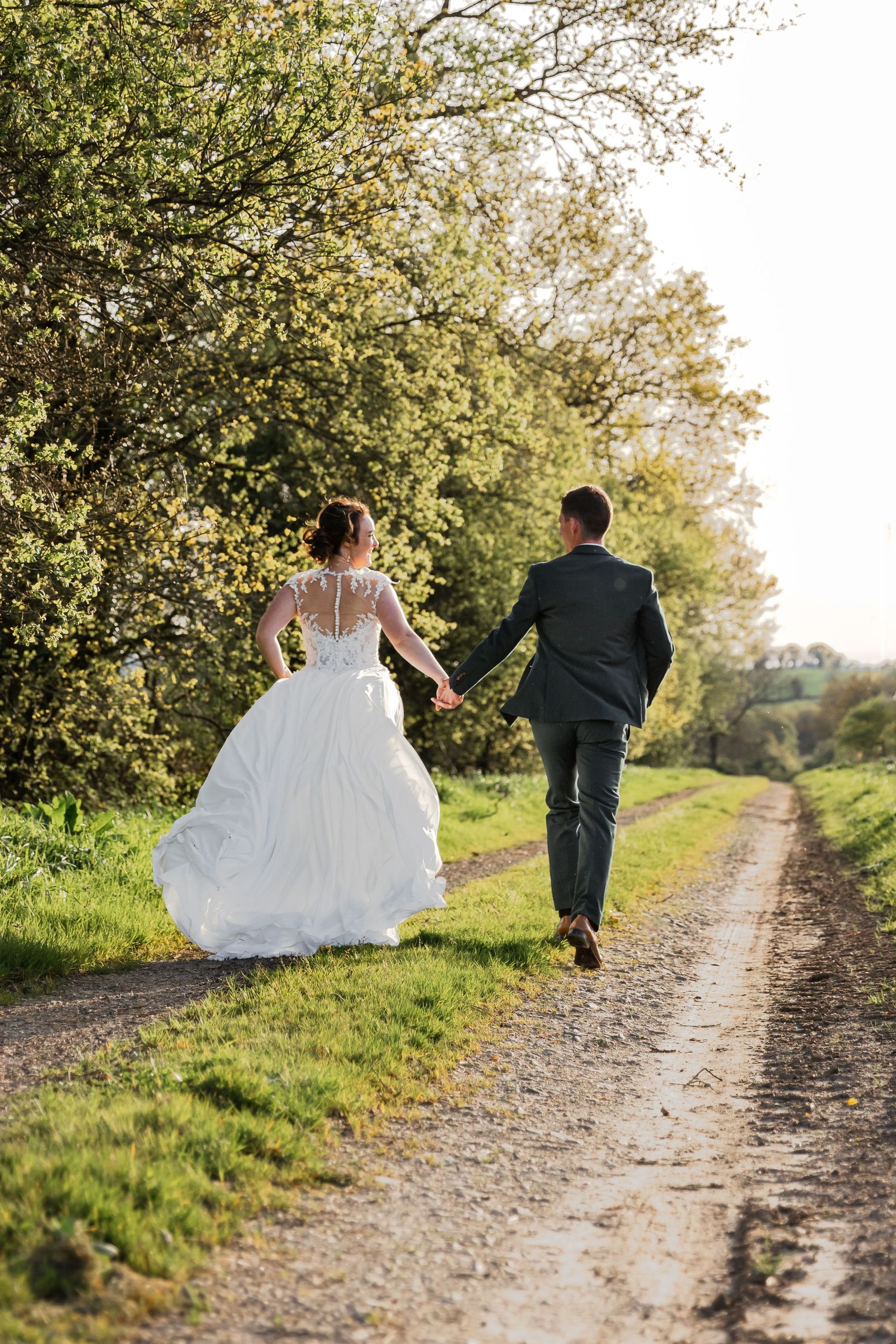 Bride and groom walking hand in hand down a scenic country path, surrounded by lush green trees on their wedding day.