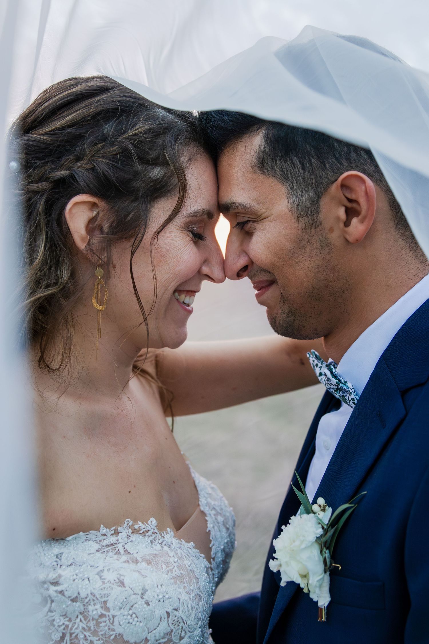 Bride and groom touching foreheads under veil, sharing romantic moment in wedding attire.