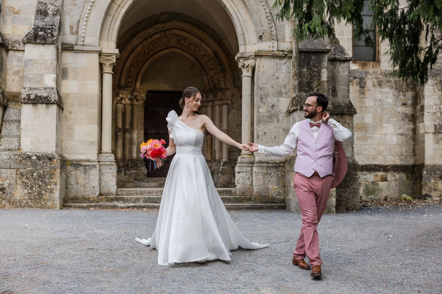 Bride in white gown and groom in pink suit hold hands outside a stone church after their wedding ceremony.