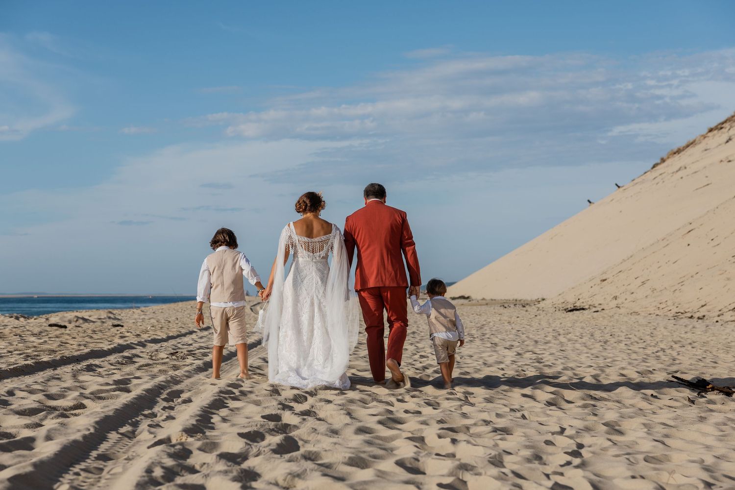 A wedding couple walks hand-in-hand with two children on a sandy beach, bride in white gown, groom in red suit.