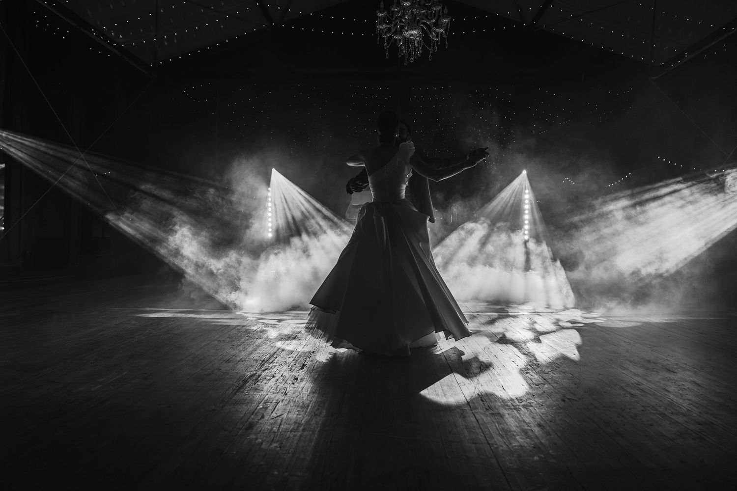 Black and white photo of a dancer performing on stage with dramatic light beams and smoke in a dark venue.