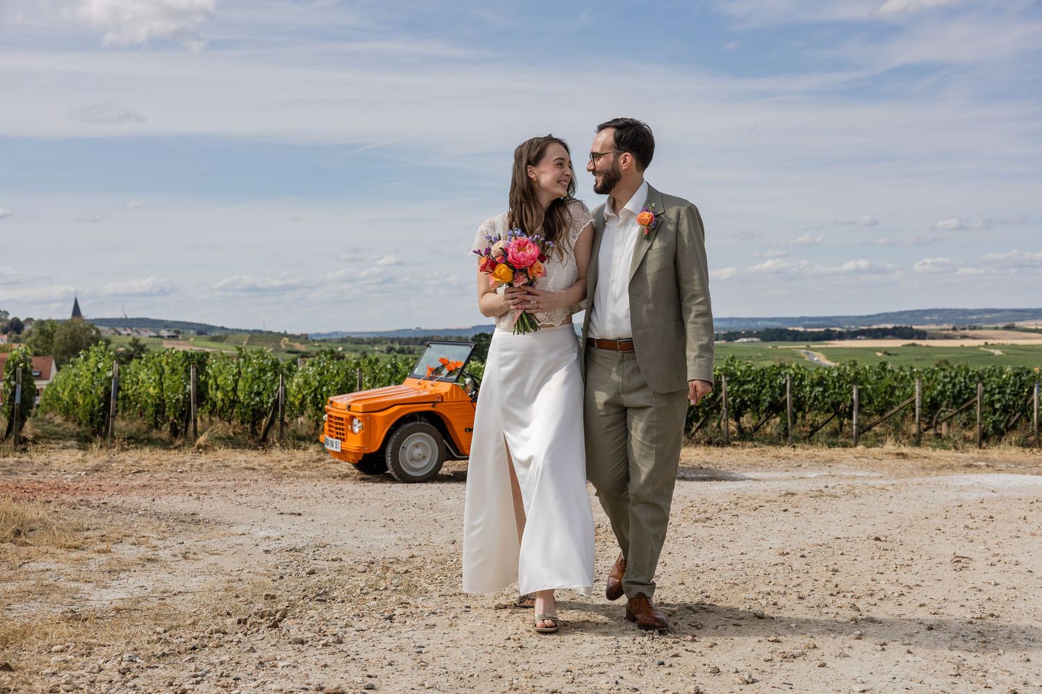 Bride and groom walking through vineyard with vintage orange car, holding colorful bouquet on sunny wedding day.