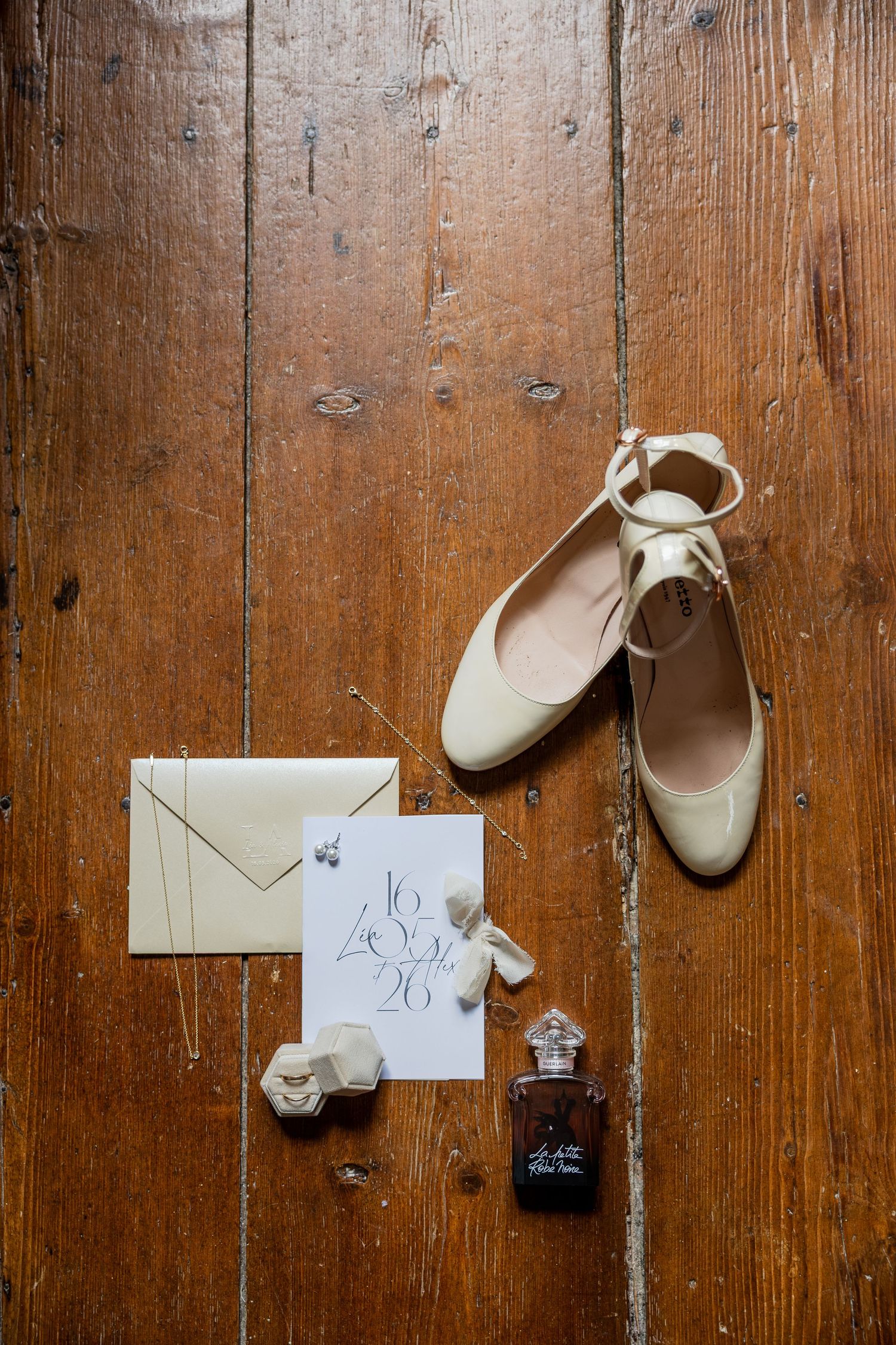 Elegant bridal flat shoes, cream envelope, wedding date card, pearl earrings, and perfume bottle on rustic wooden floor.