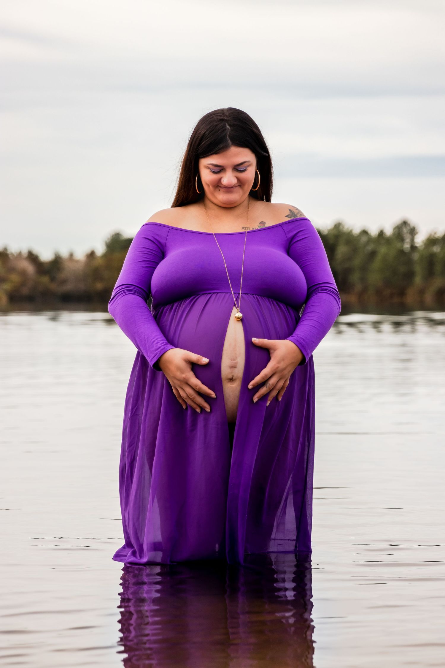 Pregnant woman in purple off-shoulder gown standing in lake water, cradling baby bump, with forest background.