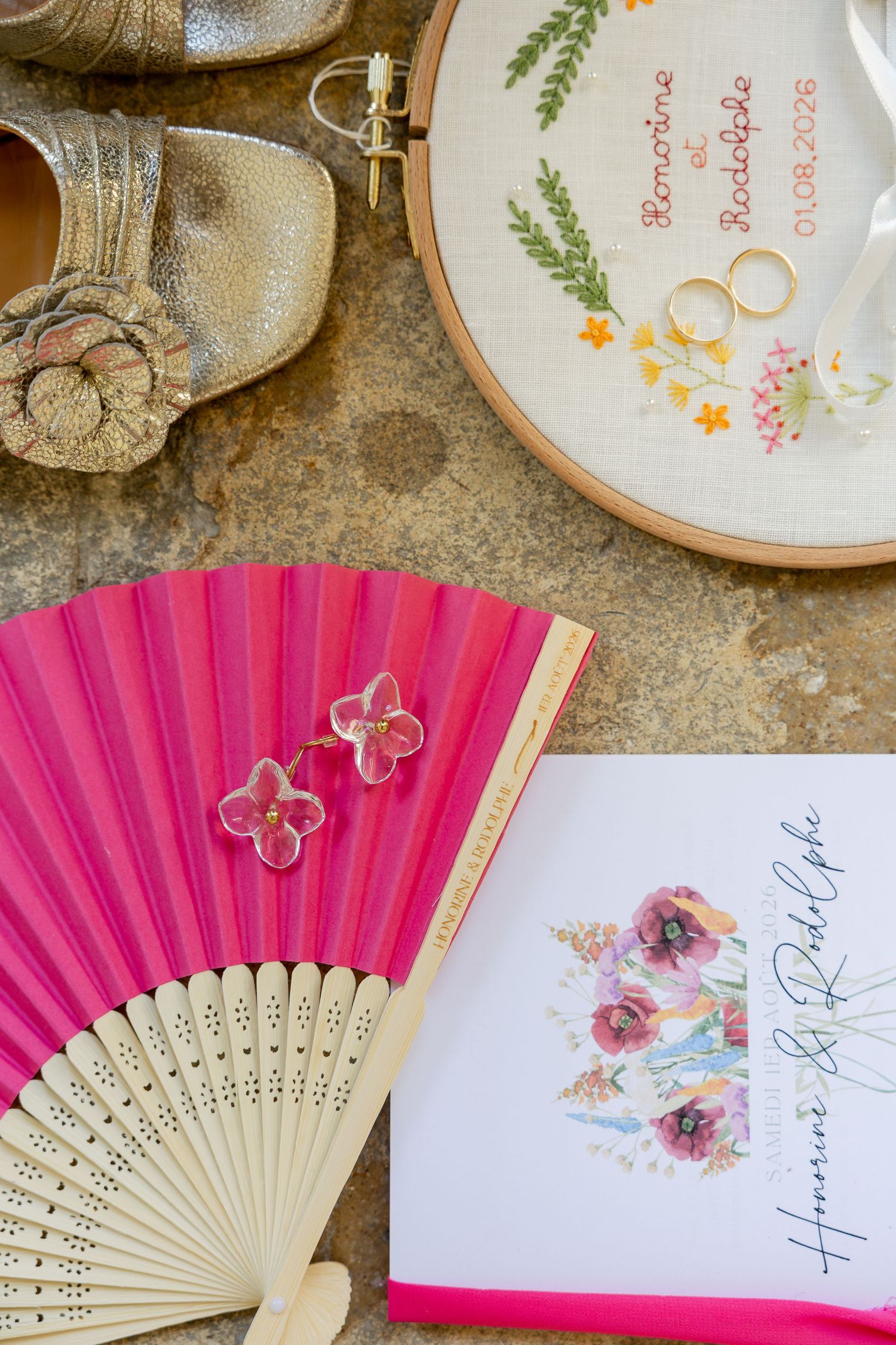 Pink fan, floral earrings, wedding rings on embroidery hoop, gold shoes, and wedding program arranged as bridal flat lay.