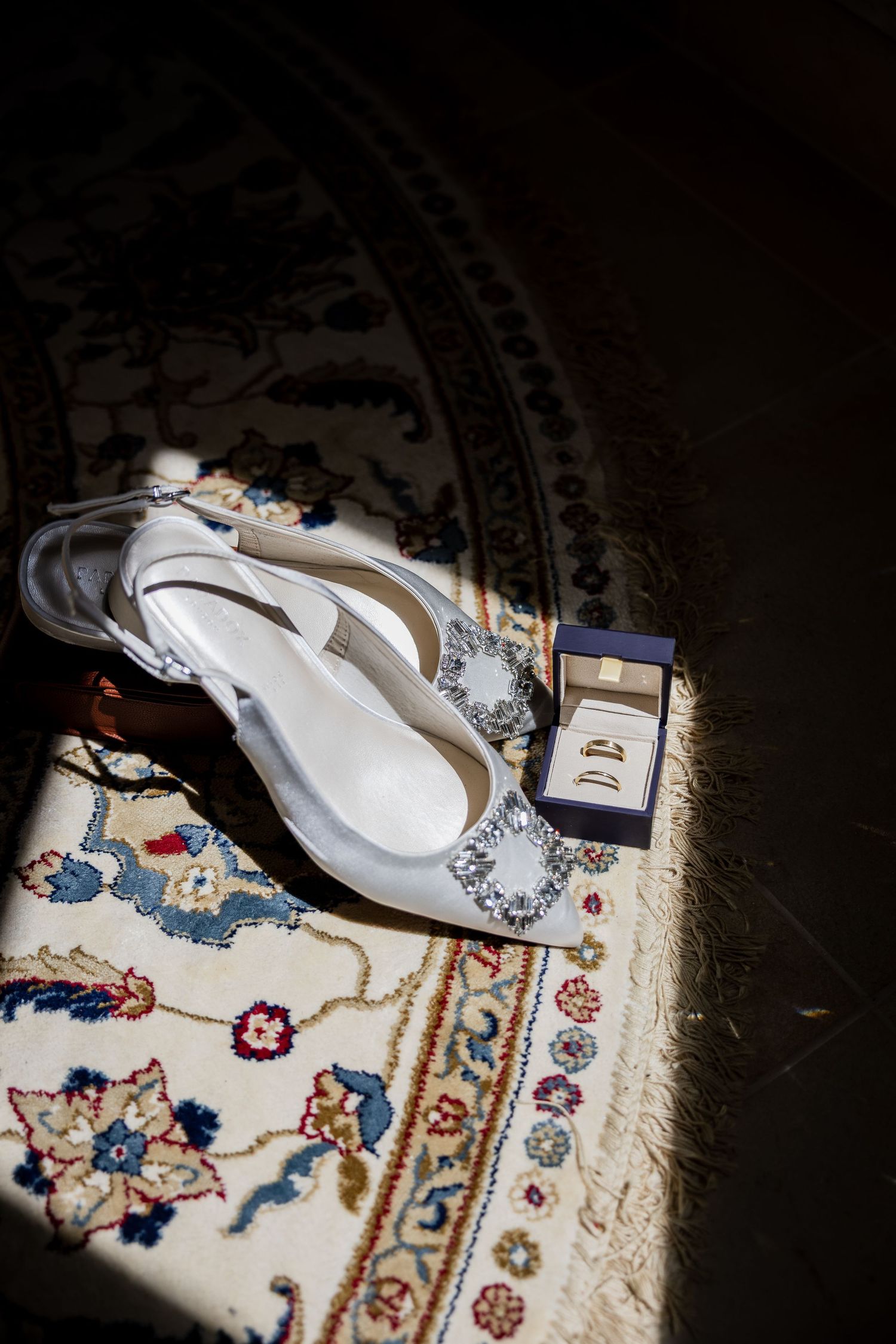White bridal heels with crystal embellishments beside a jewelry box on a floral patterned rug in soft natural light.